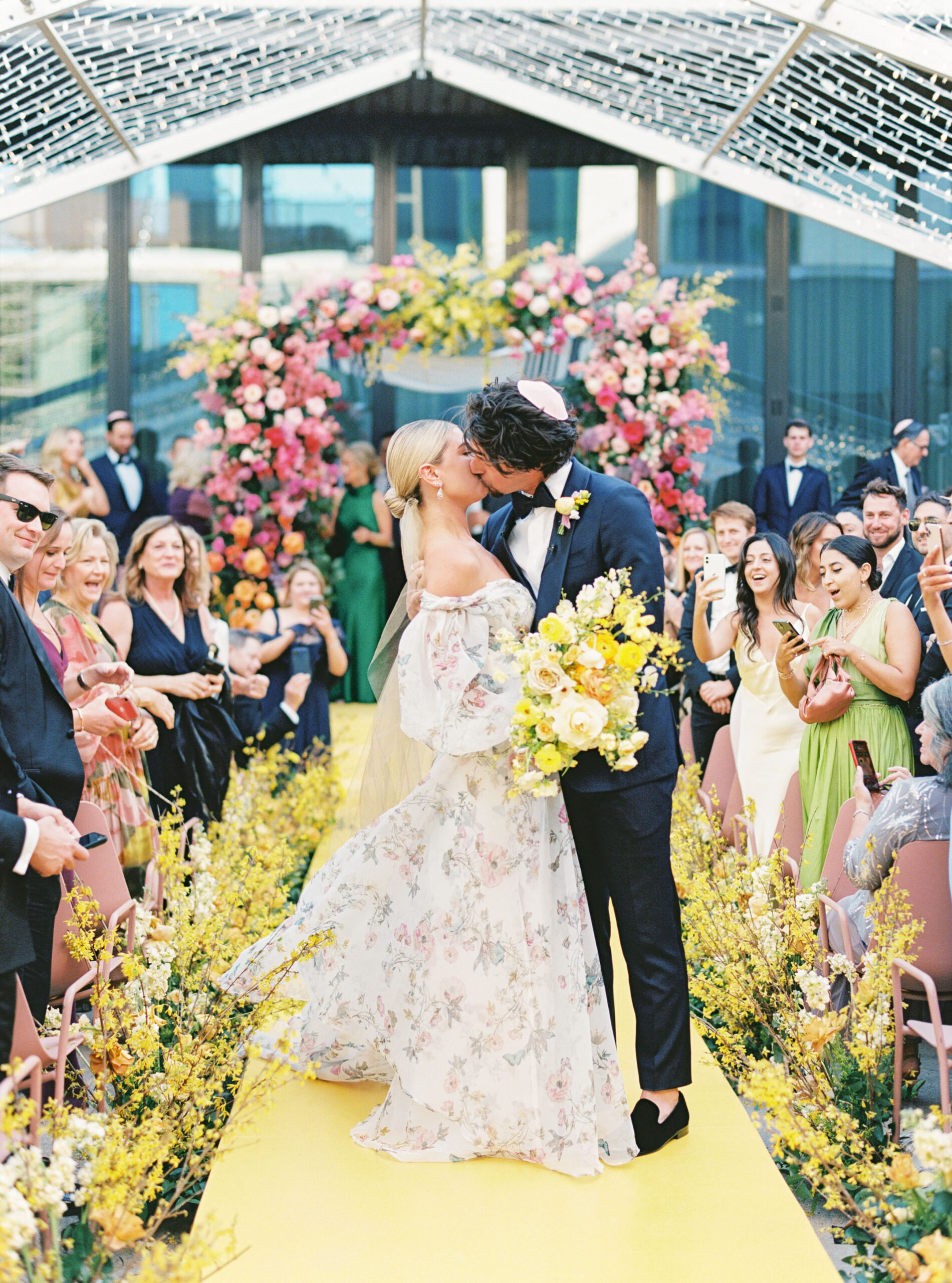 Bride and groom kissing after their ceremony, with their bright yellow aisle and colorful Chuppah behind them.